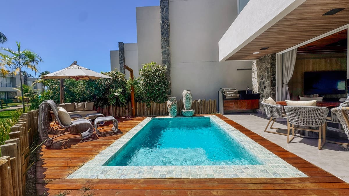 Piscina azul entre deck de madeira e varanda coberta com mesa, poltronas e sala aberta ao fundo