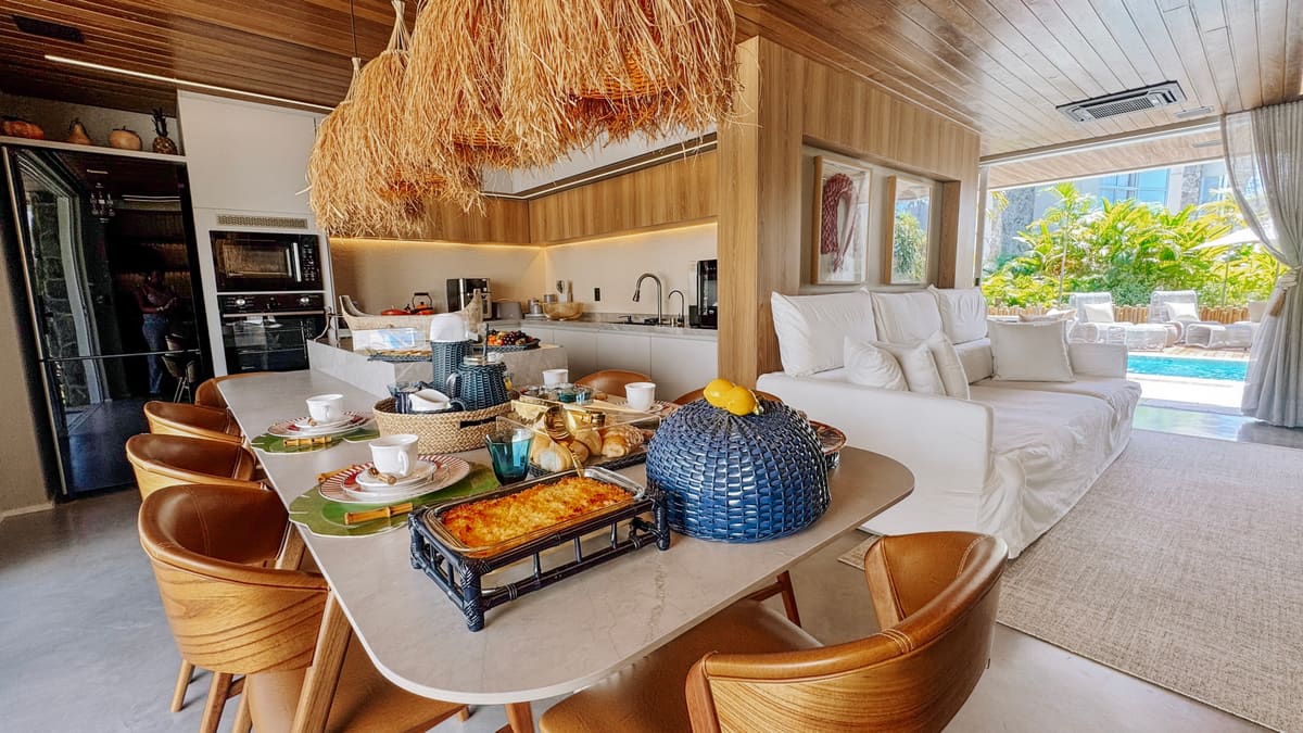 Mesa de jantar posta para café da manhã, sofá branco e porta de vidro com piscina ao fundo