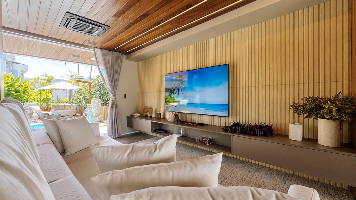 Sala de estar com sofás brancos, televisão em painel ripado e abertura para varanda com guarda-sol