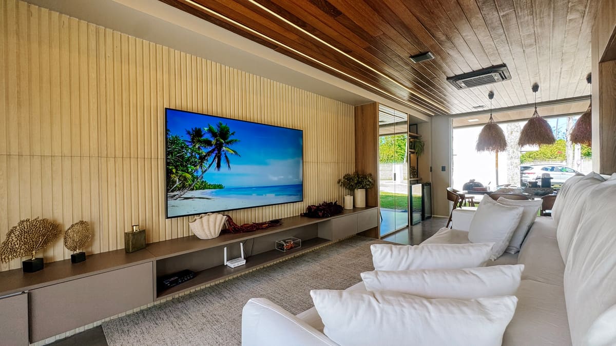 Sala com sofá branco, televisão grande em painel ripado e porta de vidro para a área externa