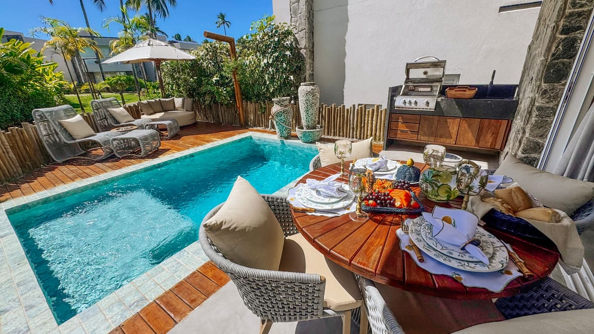 Mesa redonda de madeira posta com louça e frutas, piscina azul ao lado e churrasqueira ao fundo